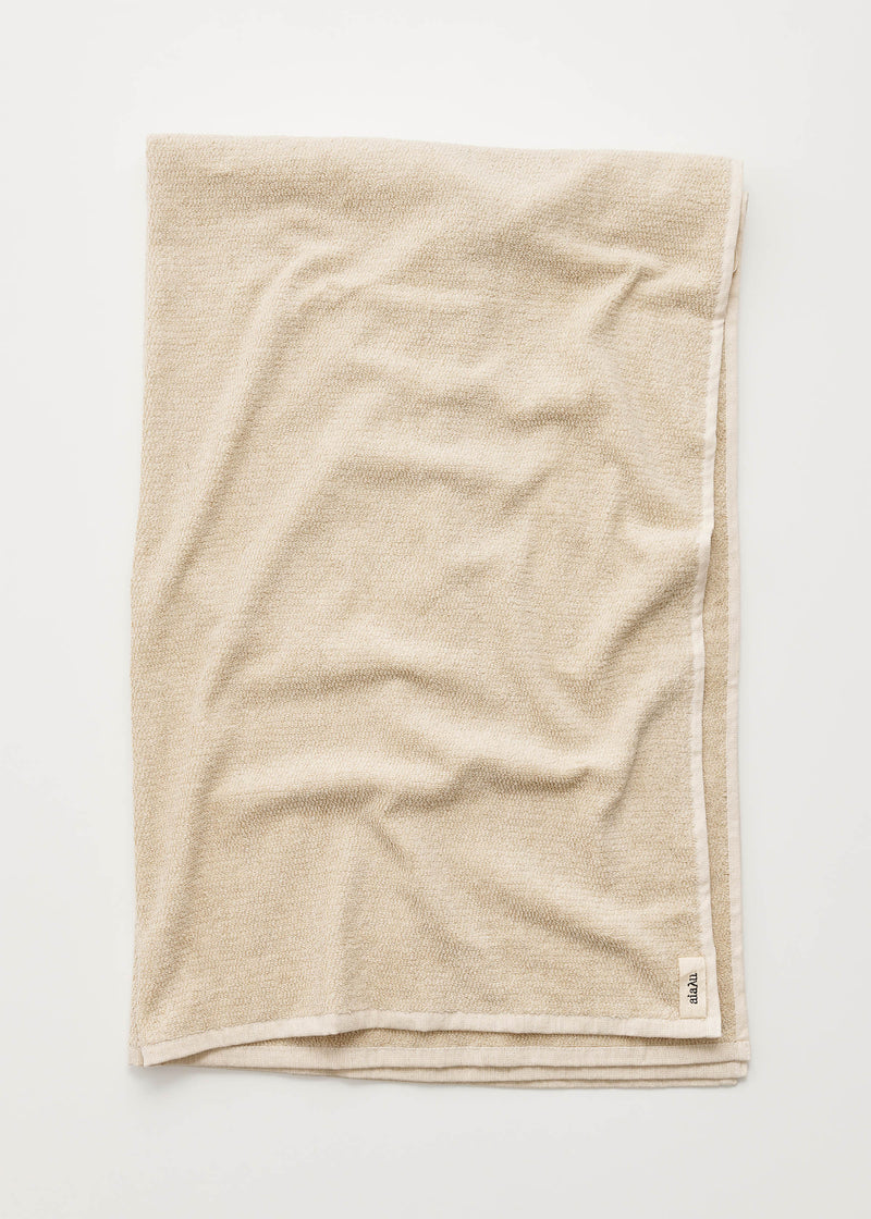 Et beigefarvet håndklæde ligger foldet på en hvid baggrund, let krøllet. En etiket med teksten "aiaaa" er synlig i nederste højre hjørne.