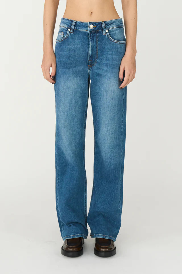 Et par mellemblå jeans bæres af en person stående foran en hvid baggrund. Bukserne er vide i benene og går ned til brune sko.