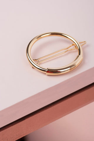 En gylden, cirkelformet hårklemme hviler på en overflade, der er opdelt i lyserøde og rosaguld segmenter.