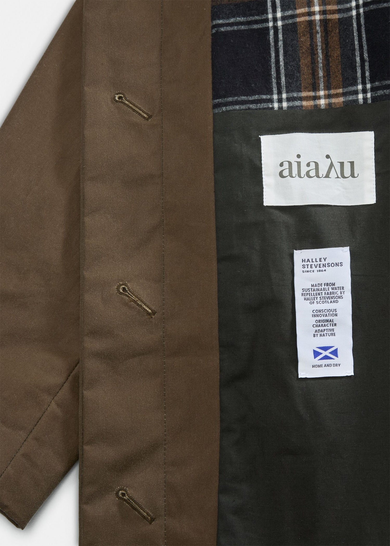 En brun frakke viser sin indre foring, som har tekstetiketter med påskrifterne "αίαλυ", "HALLEY STEVENSONS SINCE 1864", "MADE FROM SUSTAINABLE WATER REPELLENT FABRIC BY HALLEY STEVENSONS OF SCOTLAND", "CONSCIOUS INNOVATION ORIGINAL CHARACTER ADAPTIVE BY NATURE" og "HOME AND DRY".