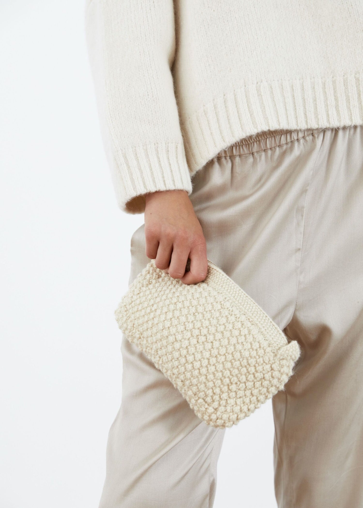 En person holder en cremefarvet, strikket pung. Pungen er holdt mod en baggrund af en hvid sweater og silkebukser.