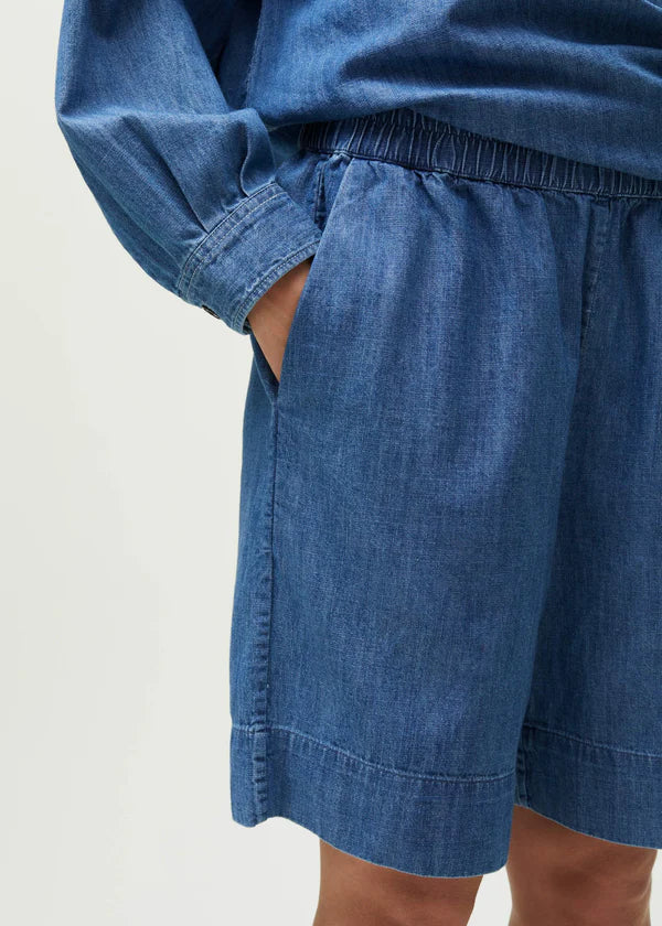 En person iført en denim-top og shorts med elastisk talje, står foran en hvid baggrund. En hånd er delvist i lommen på shortsene.