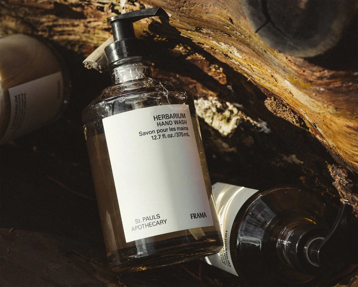 En flaske med pumpe indeholdende "HERBARIUM HAND WASH, Savon pour les mains 12.7 fl. oz./375mL, St. PAULS APOTHECARY, FRAMA" ligger blandt andre flasker og træstykker.