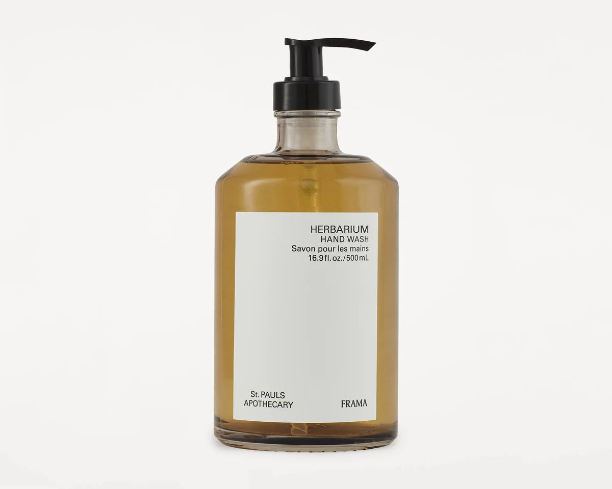 En klar glasflaske med en sort pumpe indeholder flydende håndsæbe, placeret på en hvid baggrund. Etiketten læser: "HERBARIUM HAND WASH Savon pour les mains 16.9 fl. oz./500mL, St. PAULS APOTHECARY, FRAMA".