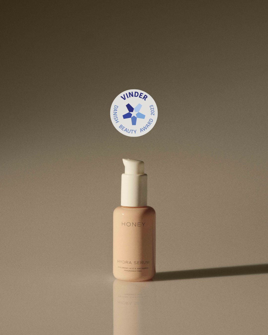 En flaske med "HONEY HYDRA SERUM" (HYALURONIC ACID & NIACINAMIDE FRAGRANCE FREE) står på en overflade. Over flasken er et mærke med teksten "VINDER DANISH BEAUTY AWARD 2023".