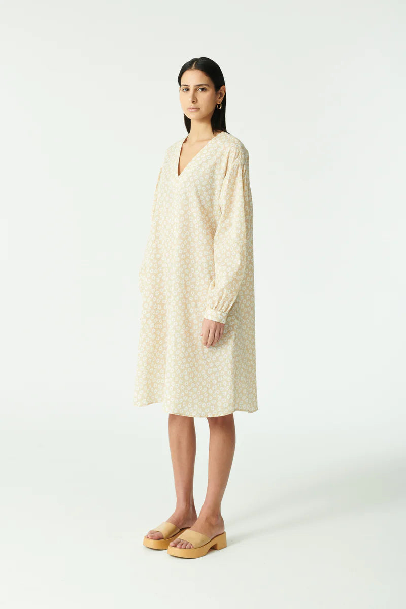 En kvinde poserer i en beige blomstret kjole med lange ærmer og V-hals. Hun har sandaler på og står på en hvid baggrund.