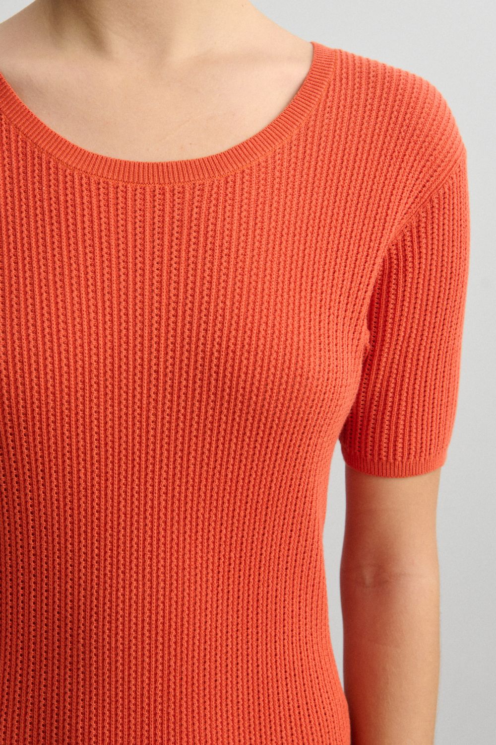 En kvinde bærer en orange, strikket bluse med korte ærmer. Blusen har en rund halsudskæring og er vist mod en lys, neutral baggrund.