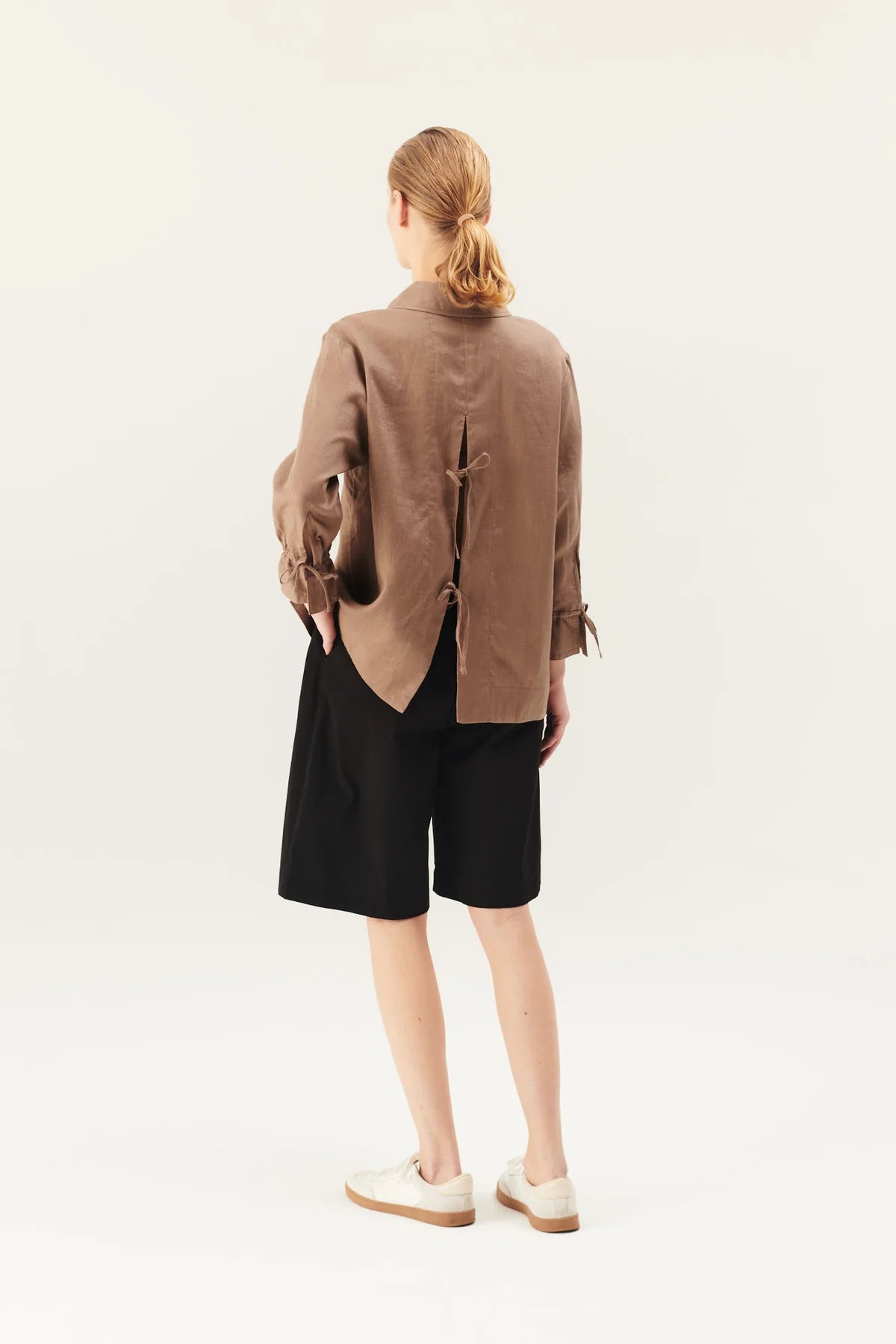 En kvinde, set bagfra, står i et studiemiljø. Hun bærer en brun skjorte med snøre bagpå, sorte shorts og hvide sneakers. Hun har den ene hånd i lommen.