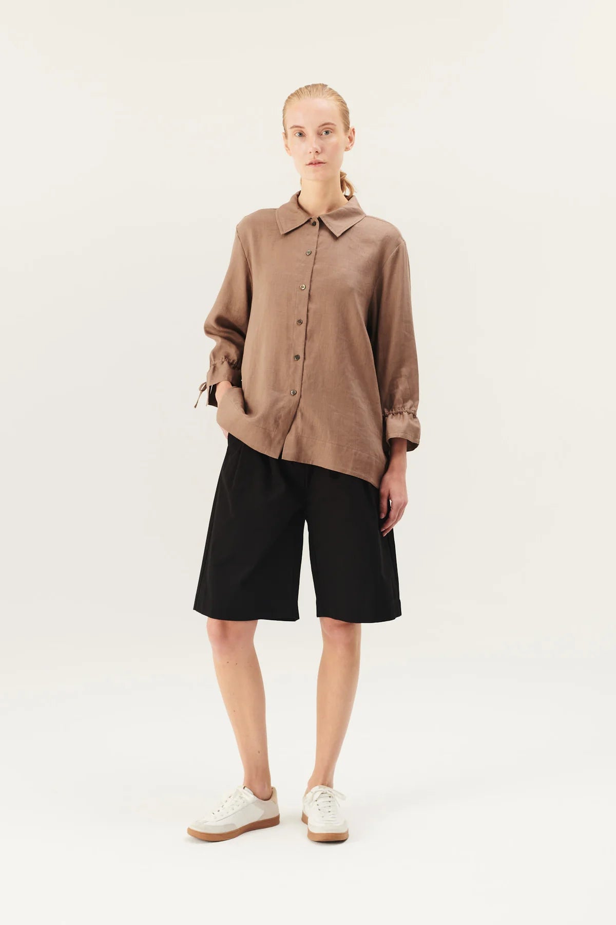 En kvinde står, iført en brun linnedskjorte, sorte shorts og hvide sko, med en hånd i lommen, foran en hvid baggrund.